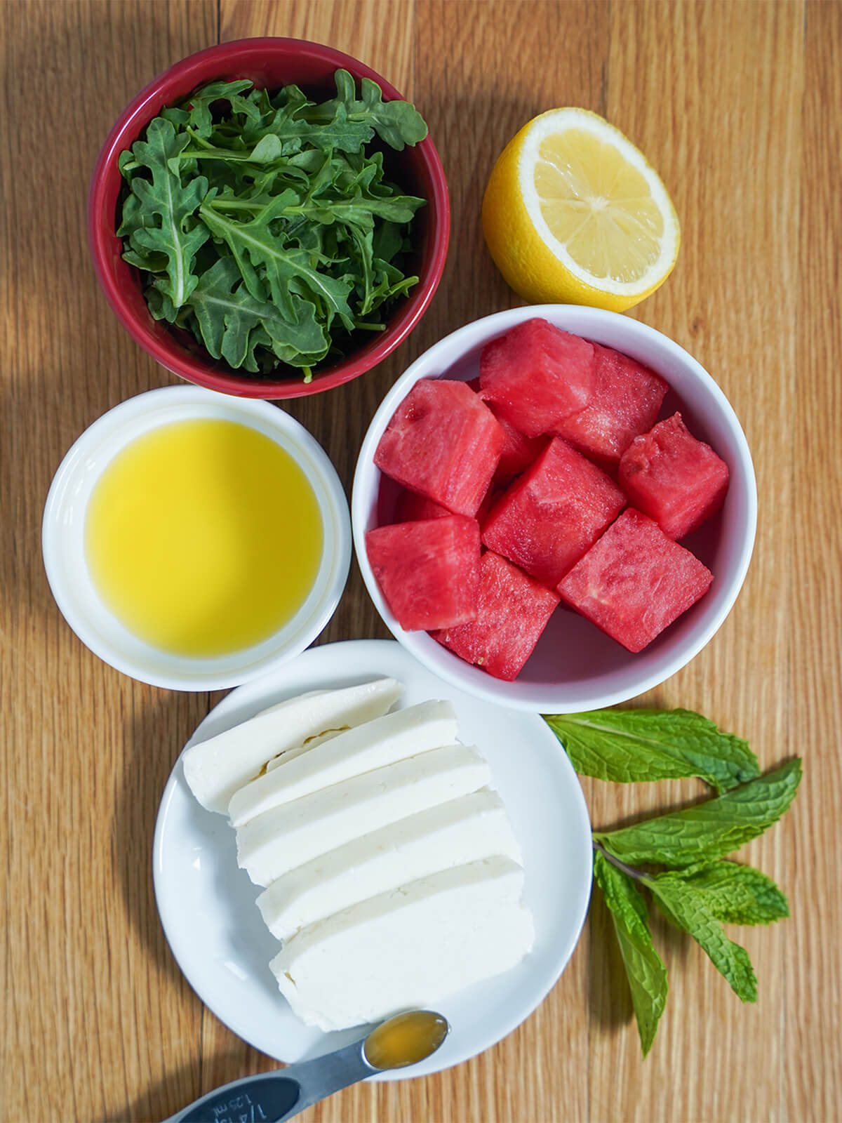 halloumi and watermelon salad ingredients bowls of diced watermelon, arugula, olive oil, sliced halloumi with part lemon and sprig of mint to side.