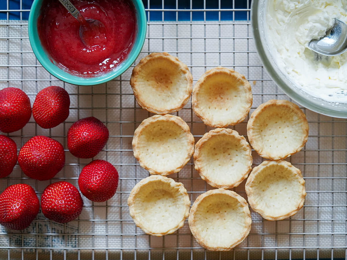 components to assemble strawberry tartlets mini pastry cases, strawberries and part view of bowl of whipped cream and bowl of strawberry glaze.