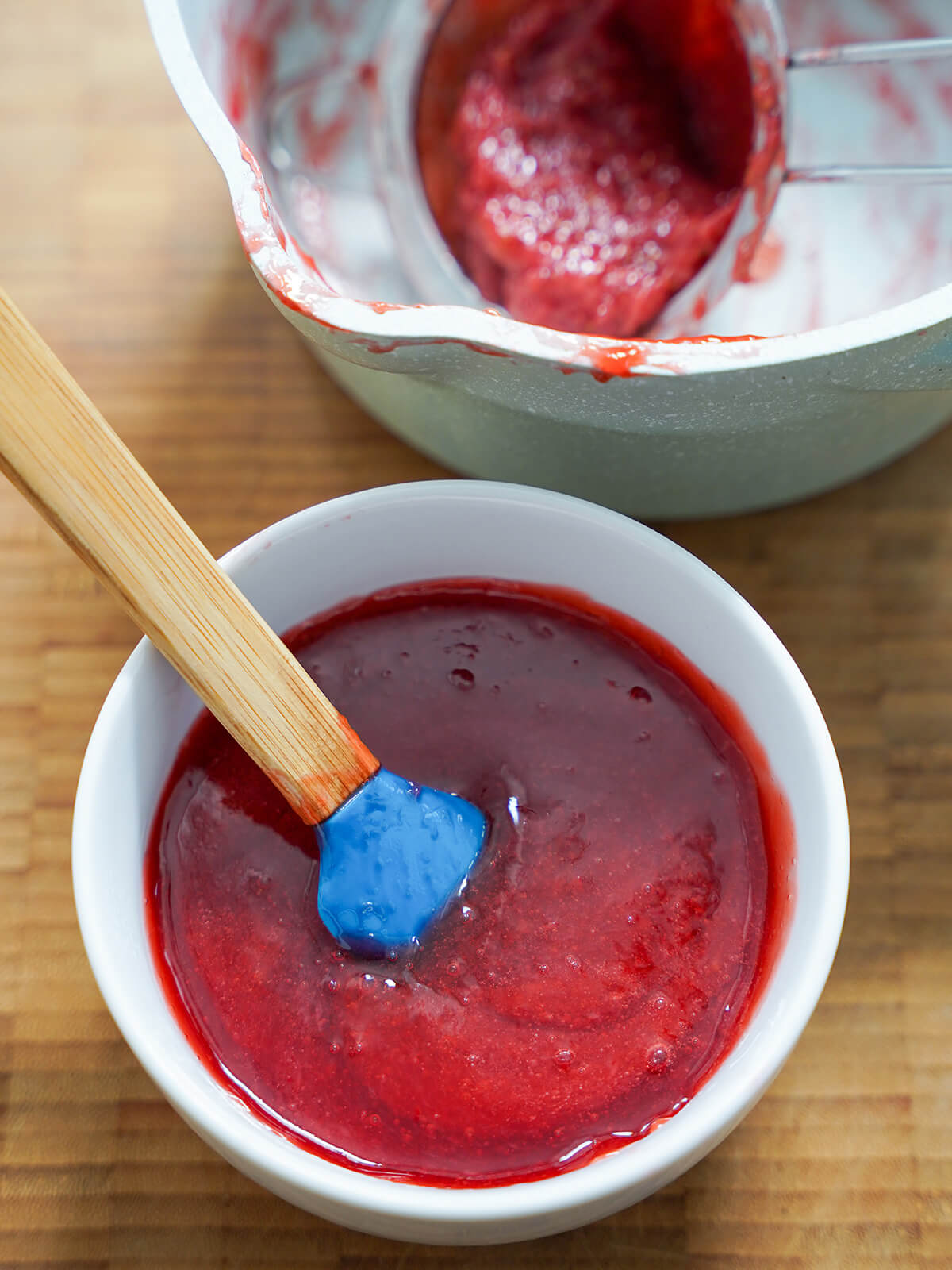 glaze for strawberry tartlets bowl of strawberry glaze with spatula in bowl and pan with strainer with strawberry solids behind.