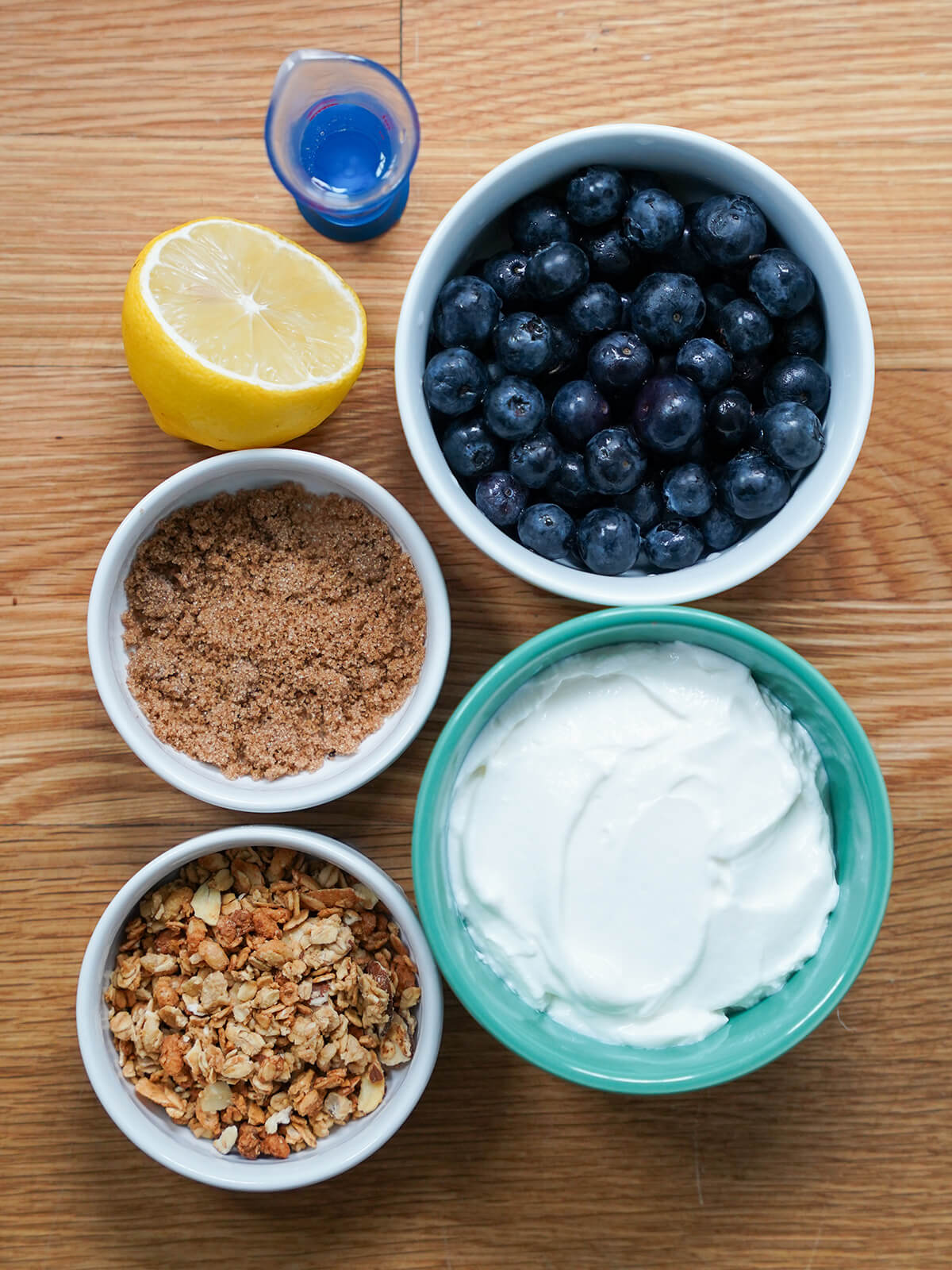 ingredients for blueberry parfait bowls of blueberries, yogurt, brown sugar, granola and part lemon from overhead.