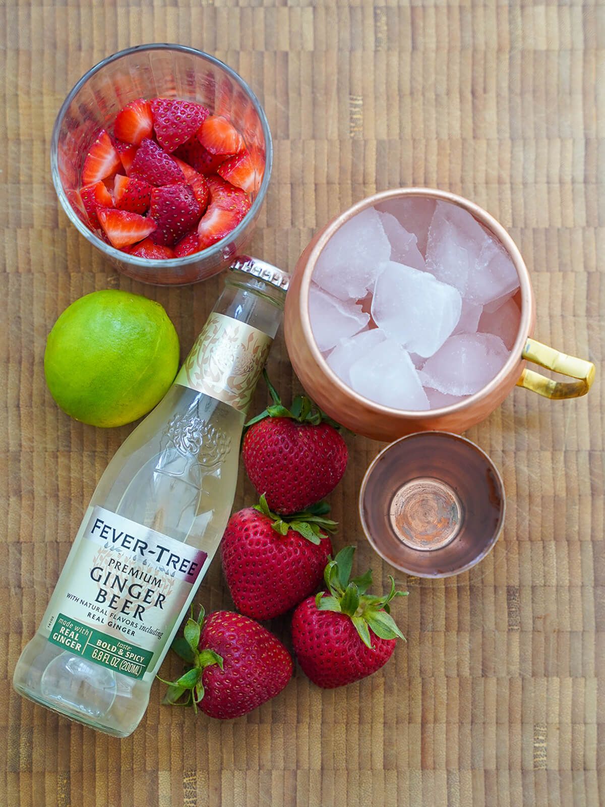 ingredients for strawberry Moscow mule bottle of ginger beer, copper mug filled with ice, strawberries, lime, measure.