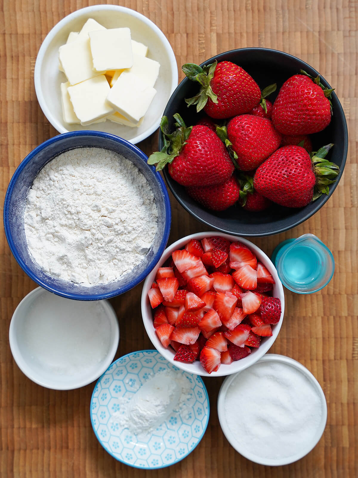 ingredients for strawberry tartlets bowls of whole and cut strawberries, bowl of flour, dishes with sugar, butter and corn starch from overhead.