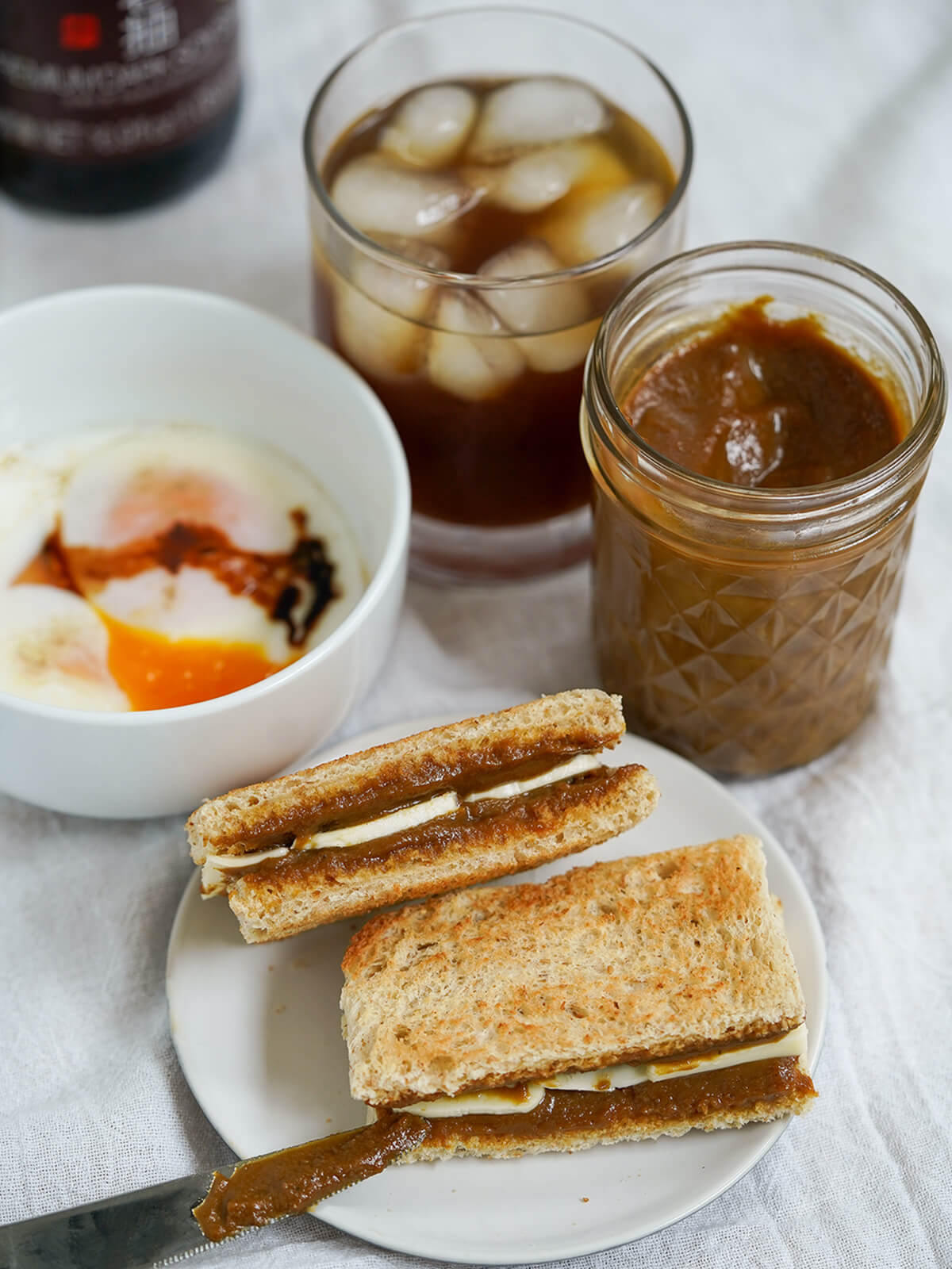 piece of kaya toast resting or corner of another with bowl of soft boiled eggs, jar of jam and glass of iced coffee behind.