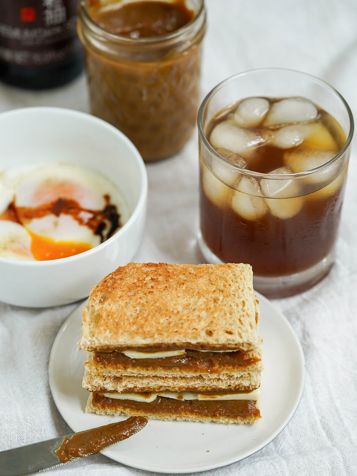 stack of kaya toast with glass of iced coffee and bowl of soft boiled eggs behind it, jar of kaya jam in background.