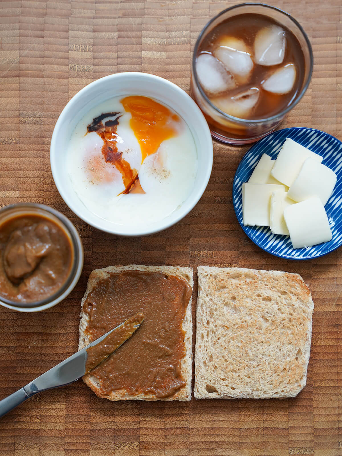 spreading kaya jam on piece of toast, bowl of eggs, plate with slices of butter and glass of coffee all from overhead.