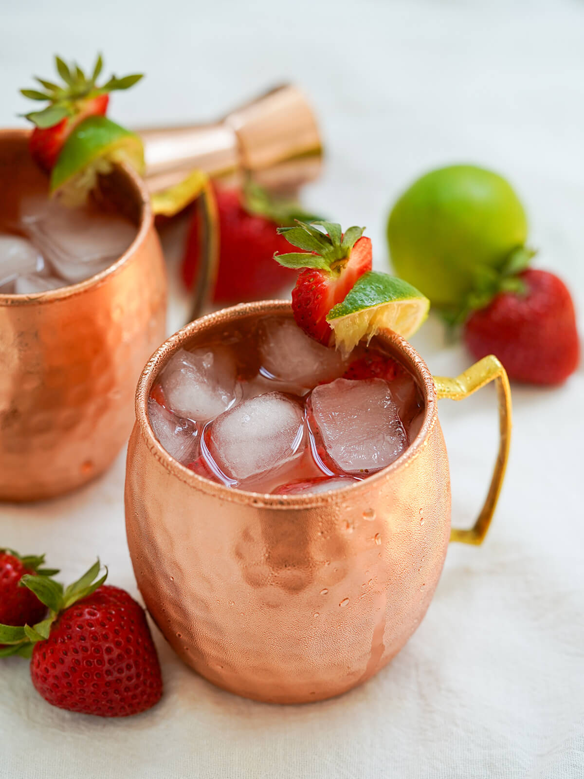 This strawberry Moscow mule is a gently fruity twist on the classic vodka ginger beer cocktail. It's easy to make, refreshing and great for summer sipping. side view of copper mug of strawberry Moscow mule with strawberry to side and more behind.