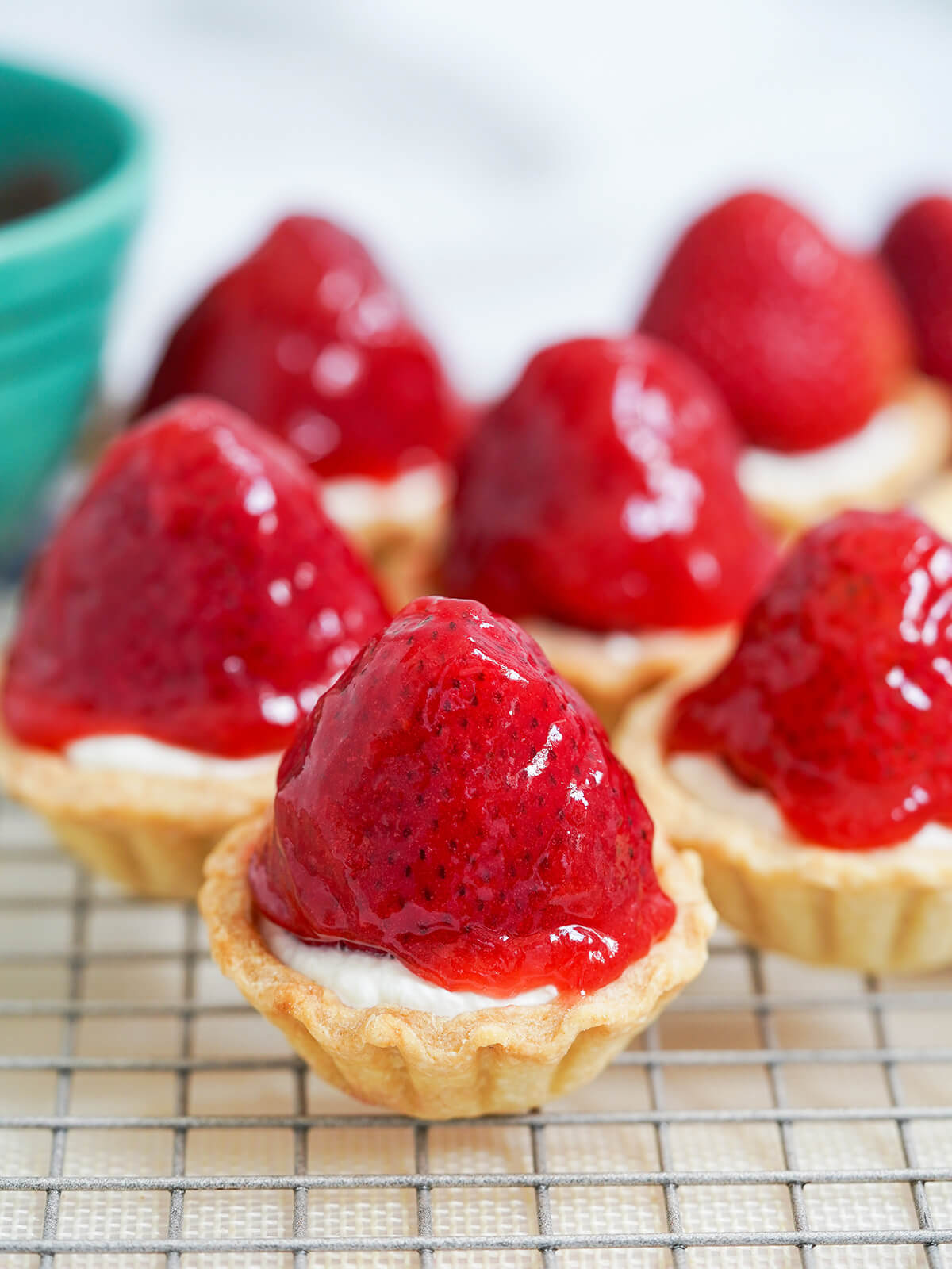 These strawberry tartlets are delicious little bites combining crisp pastry, smooth cream, juicy berries and a sweet fruity glaze. A yummy treat - maybe messy but so worth it. rows of strawberry tartlets on rack.