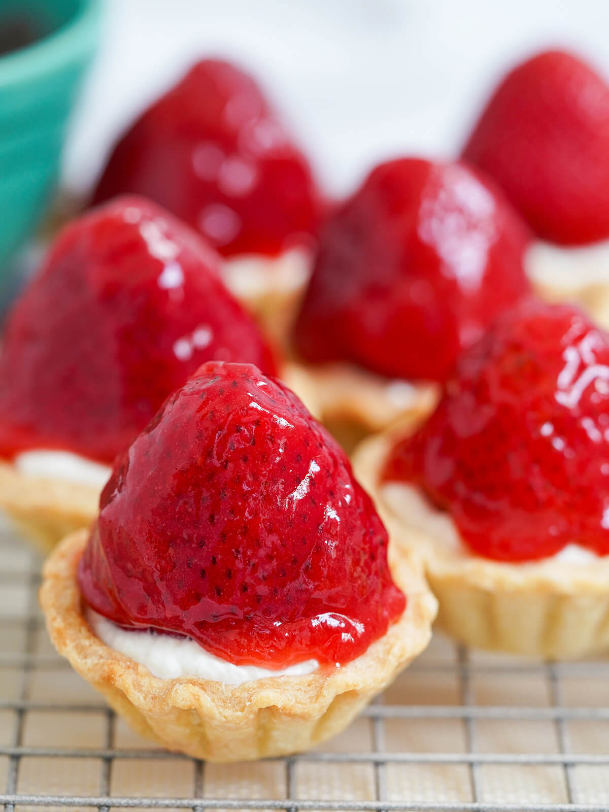 These strawberry tartlets are delicious little bites combining crisp pastry, smooth cream, juicy berries and a sweet fruity glaze. A yummy treat - maybe messy but so worth it. a few strawberry tartlets on rack showing glaze over top.