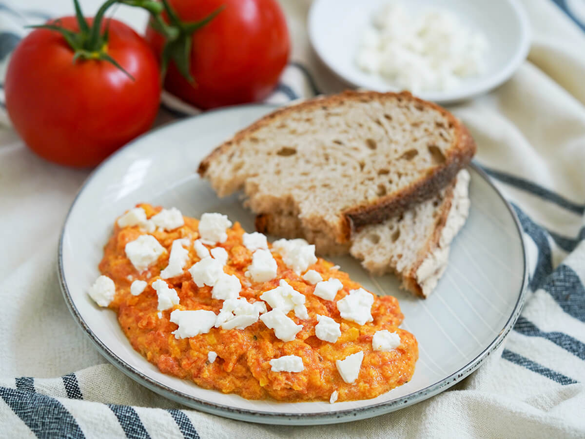 Greek eggs with tomato, strapatsada, and bread on plate with tomatoes behind.