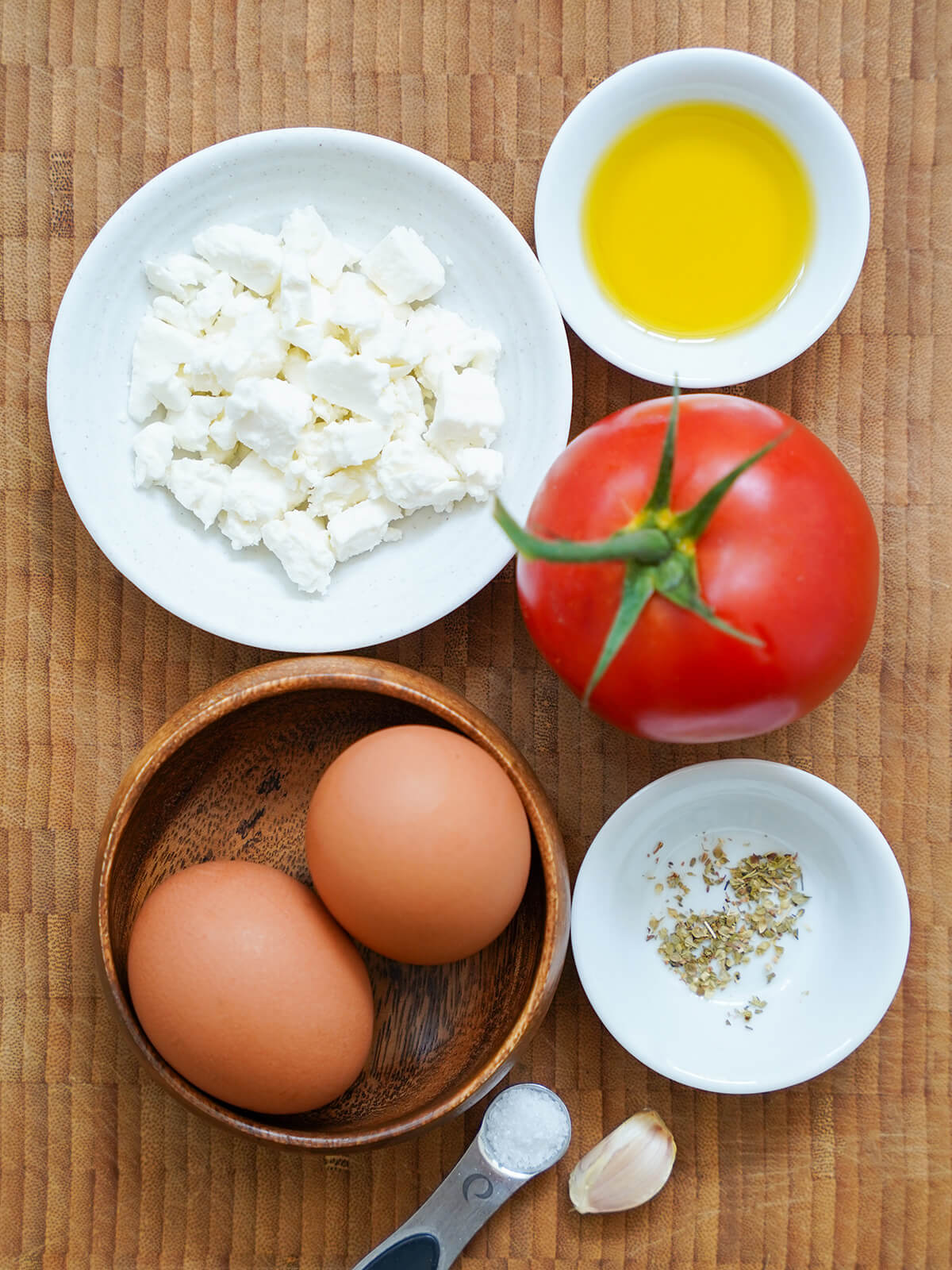 dish with feta, eggs in wooden bowl, tomato, dish with olive oil, garlic clove and salt.
