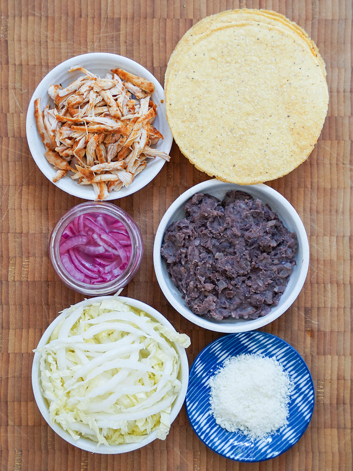 pile of corn tostadas, dishes with cooked shredded chicken, refried black beans, sliced napa cabbage, some cotija cheese and small jar of pickled red onions.