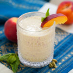 glass of peach lassi with a wedge of peach on top of glass.