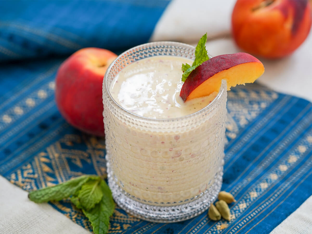 side view of glass of peach lassi with peaches in background.