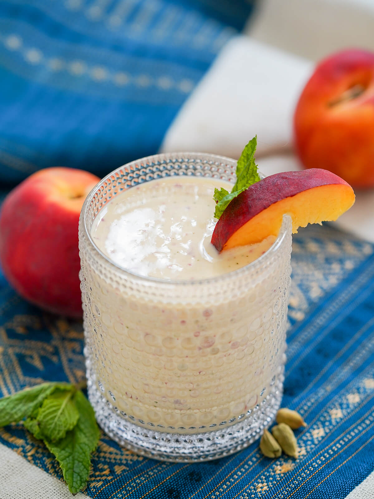 glass of peach lassi with slice of peach as garnish and part view of peaches behind.