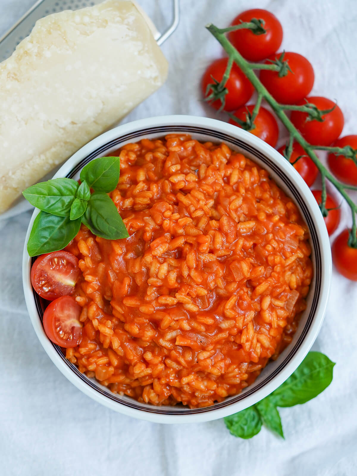bowl of tomato risotto form overhead, parmesan block and cherry tomato sprig above and basil below bowl.