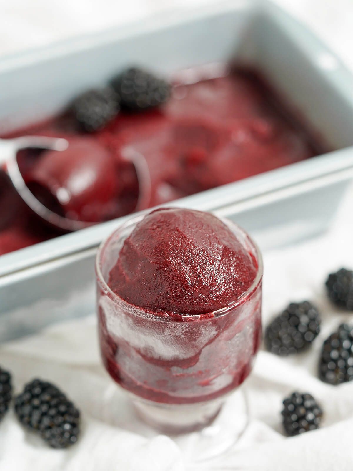 glass of blackberry sorbet in front of pan blurred in background and blackberries to sides.