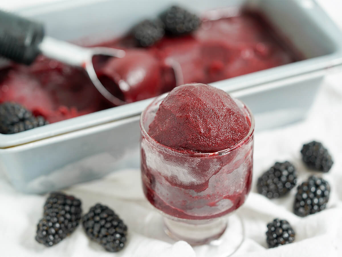 glass of blackberry sorbet in front of pan with rest and blackberries to either side of glass.