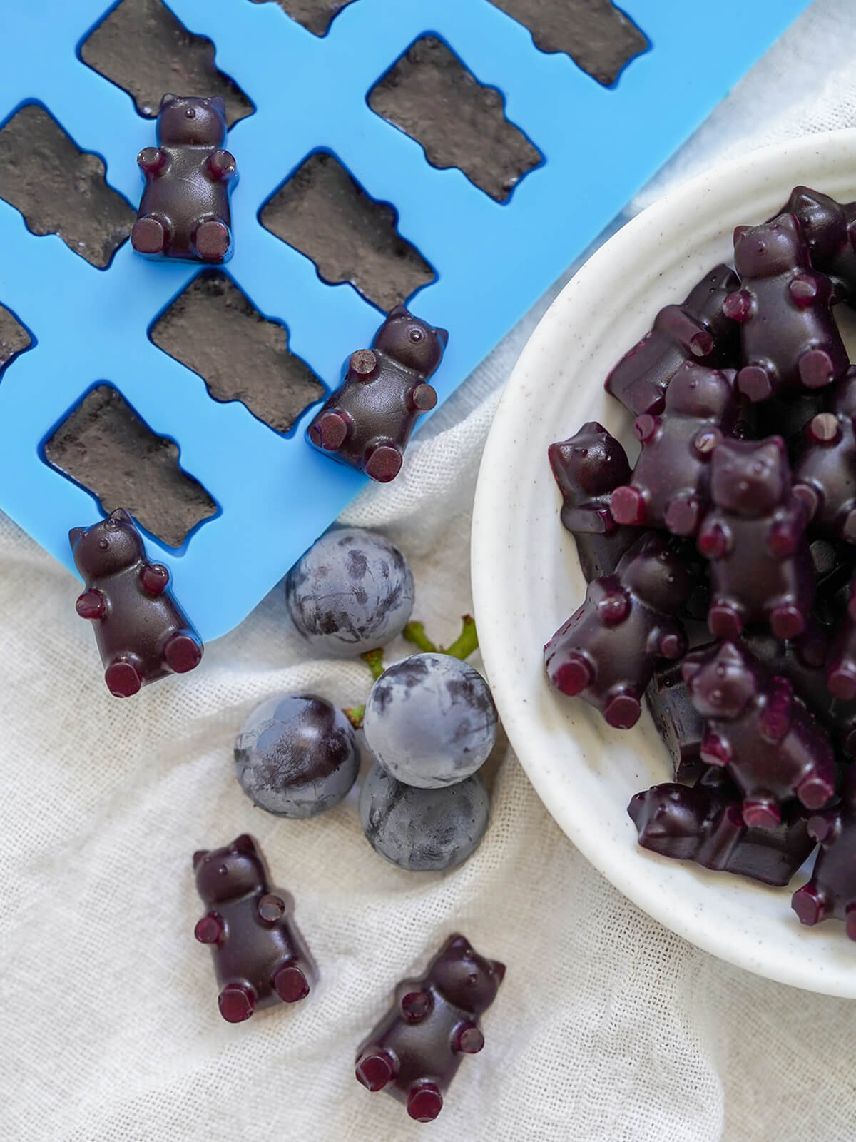 overhead view of some gummy bears on mould filled with more, a few grapes and dish with gummy bears part in view on side.