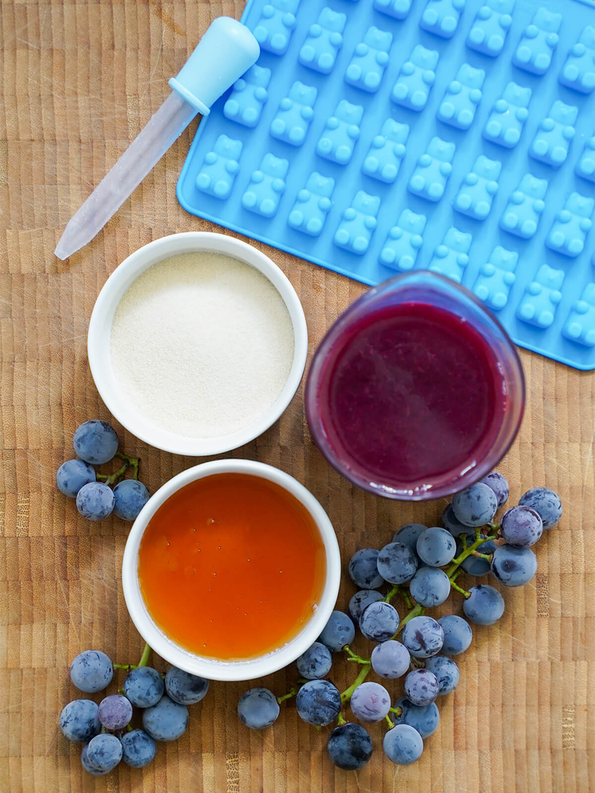 ingredients for grape gummy bears dishes with gelatin, grape juice, honey and grapes below, bear mould and dropped above.