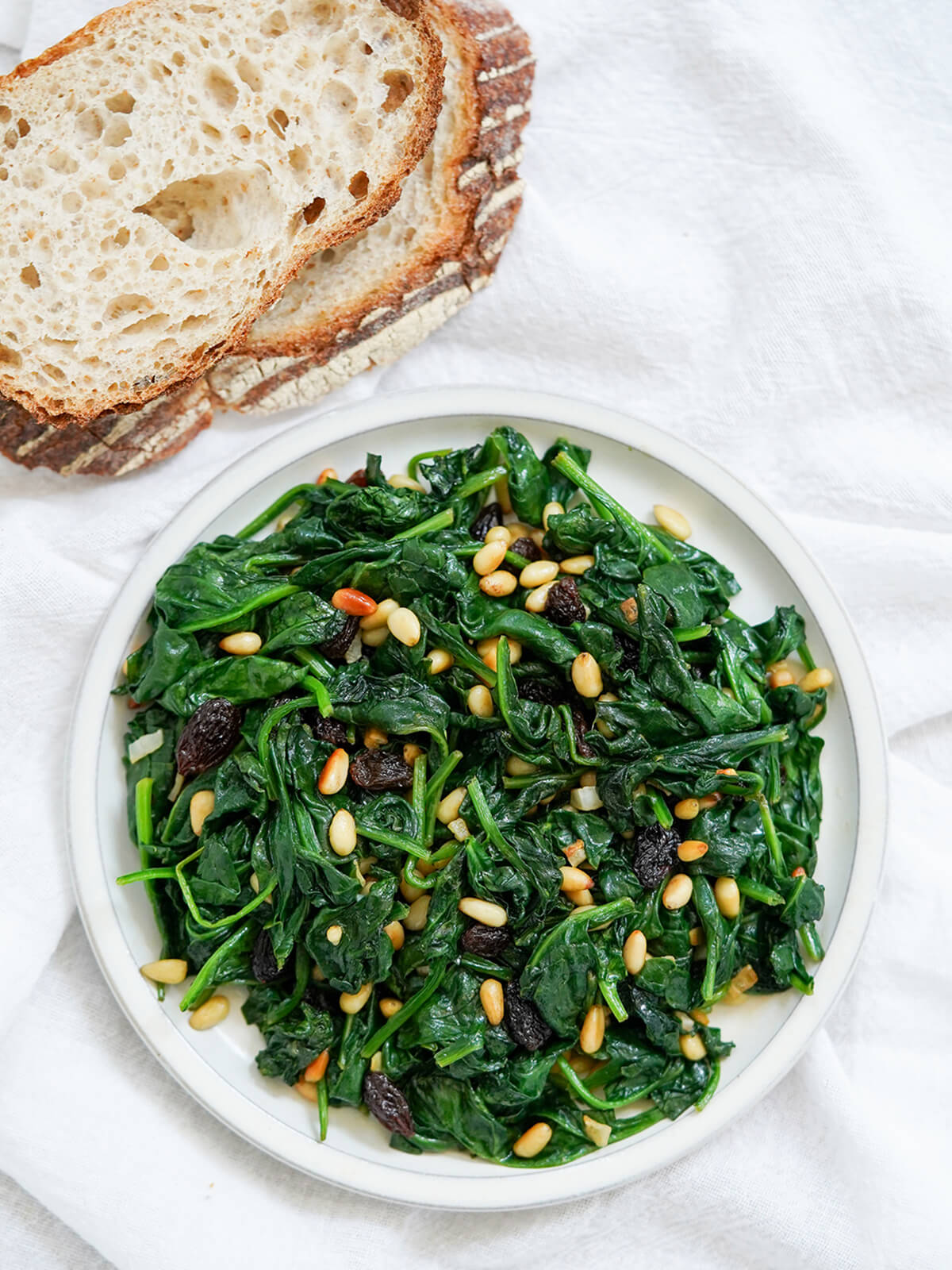 This Catalan spinach with pine nuts and raisins is wonderfully quick and easy to prepare, yet has a lovely mix of textures and flavors. Great as a tapas, side or on bread. overhead view of plate of Catalan spinach with pine nuts and raisins with bread above plate.