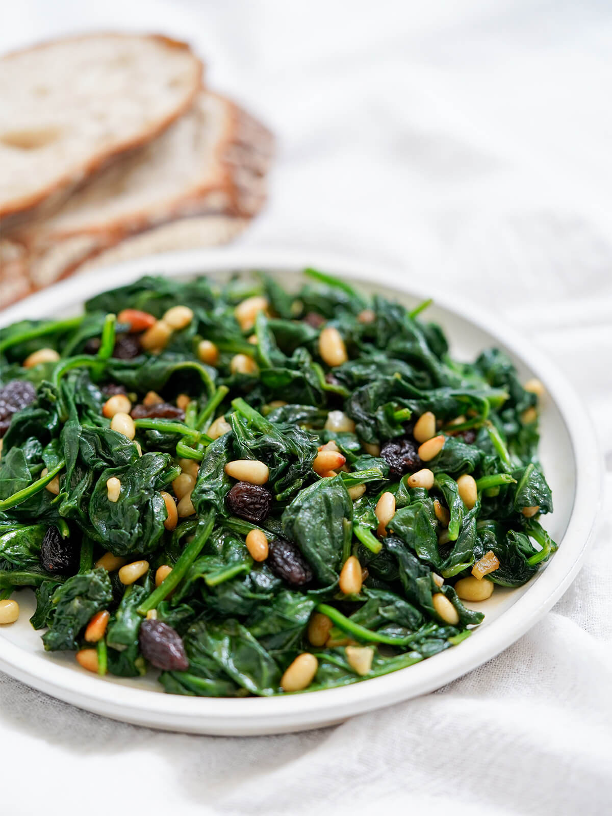 This Catalan spinach with pine nuts and raisins is wonderfully quick and easy to prepare, yet has a lovely mix of textures and flavors. Great as a tapas, side or on bread. plate of Catalan spinach with pine nuts and raisins from side with bread blurred in background.