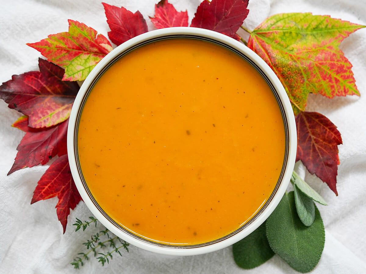 bowl of butternut squash apple soup surrounded by colorful leaves and herbs below.