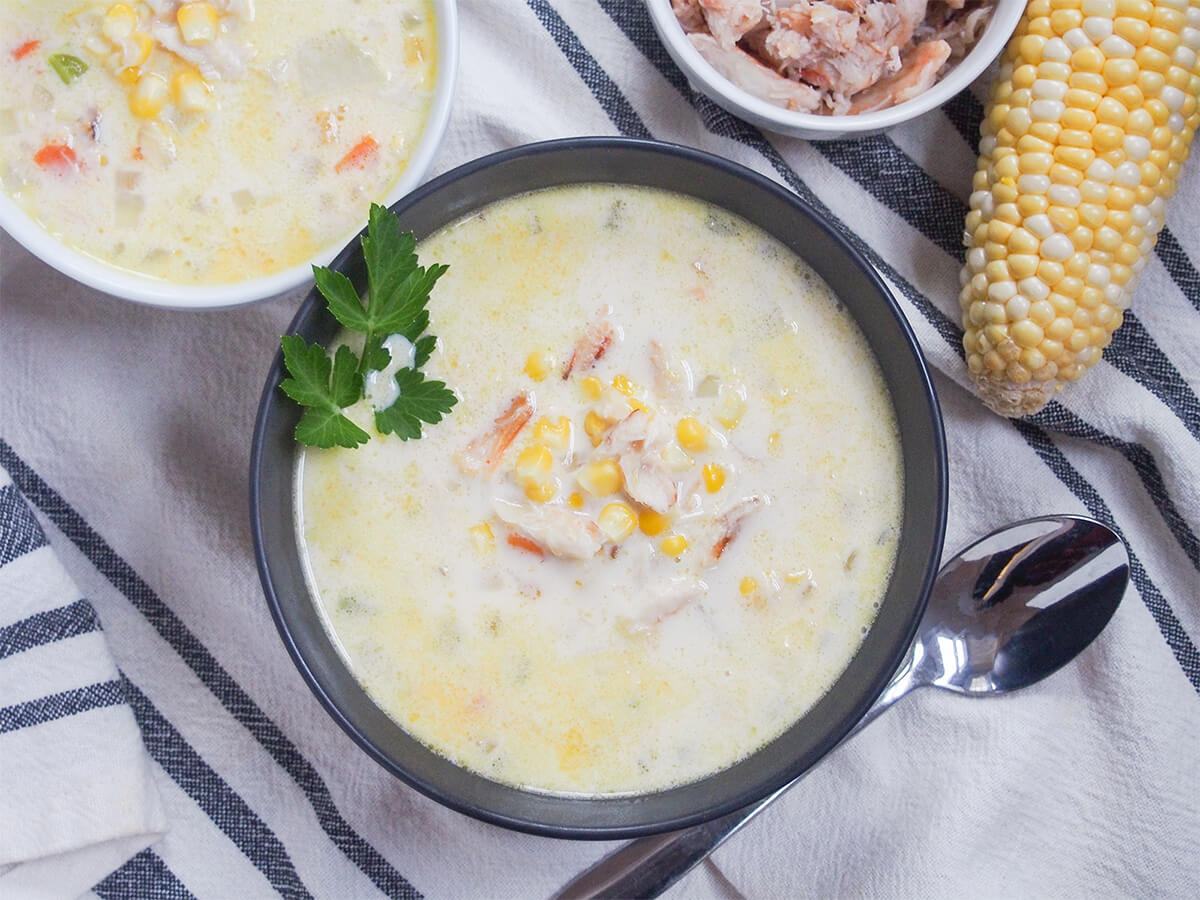 overhead view of bowl of crab and corn chowder with part corn cob to one side and part of bowl to other and spoon below bowl.