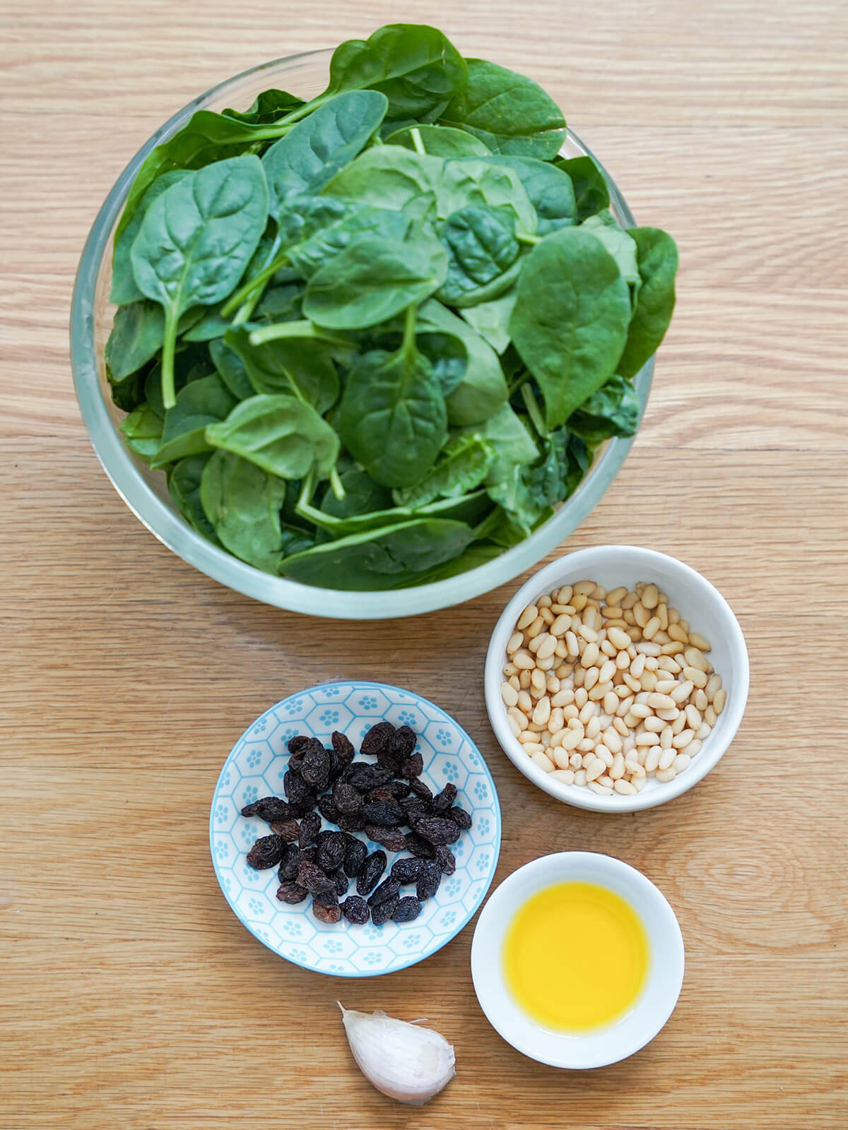 ingredients for Catalan spinach with pine nuts and raisins large bowl of spinach with dishes of raisins, pine nuts, olive oil and a garlic clove below.
