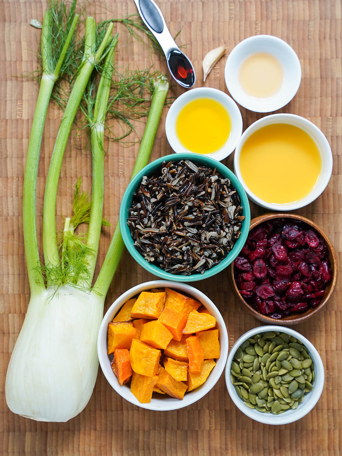bowls with cooked wild rice, squash, cranberries, pepitas with bowls of orange juice, oil, vinegar above and stem of fennel to side.