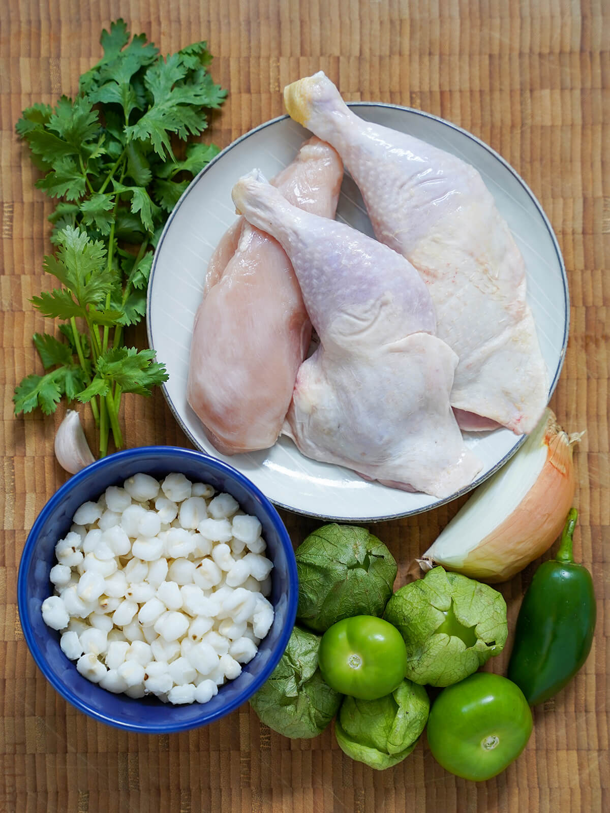 pieces of chicken on plate, bowl of hominy with tomatillos, jalepeno, wedge of onion and sprigs of cilantro around.