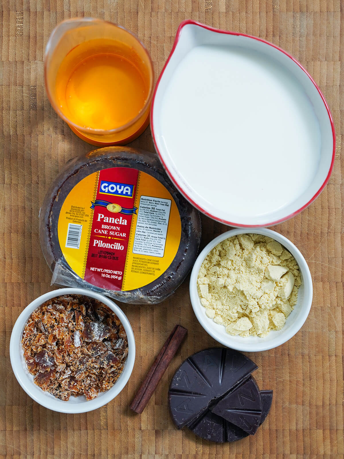 measures of milk and water, block of piloncillo with some in bowl below, bowl of masa harina and pieces of chocolate and cinnamon stick.