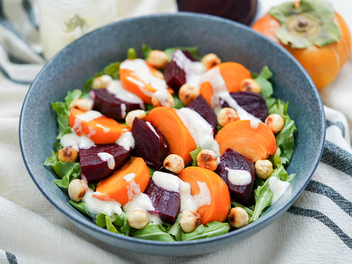 This winter beet salad is short on ingredients but full of flavor. From earthy beets to fragrant persimmon, all topped off with a delicious creamy goats cheese dressing. Great for lunch or as a side. side view of bowl of beet persimmon salad with persimmon behind bowl.