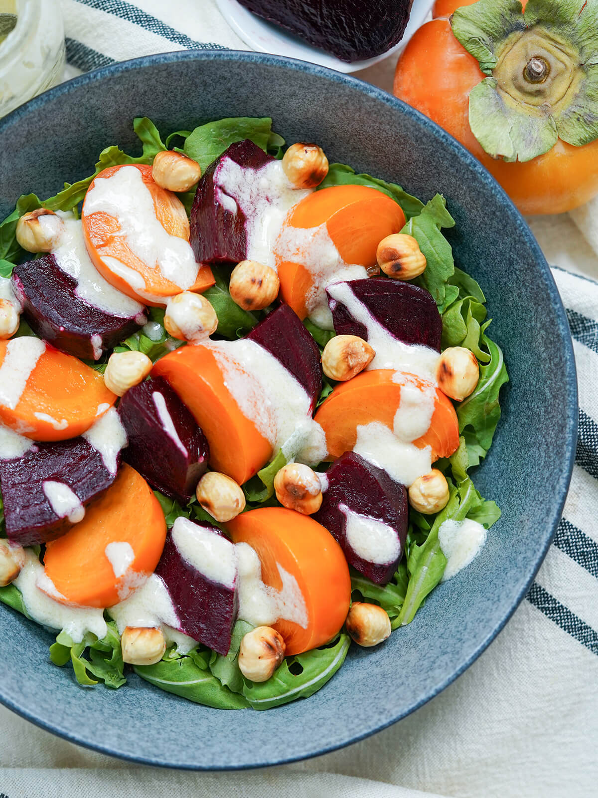 This beet persimmon salad is a simple mix of sweet and savory flavors, as well as textures, all topped off with a delicious creamy goats cheese dressing. overhead partial view of a bowl of beet persimmon salad topped with goat cheese dressing.