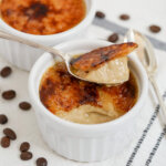 spoon of coffee creme brulee sitting on top of rest in dish with coffee beans to side.