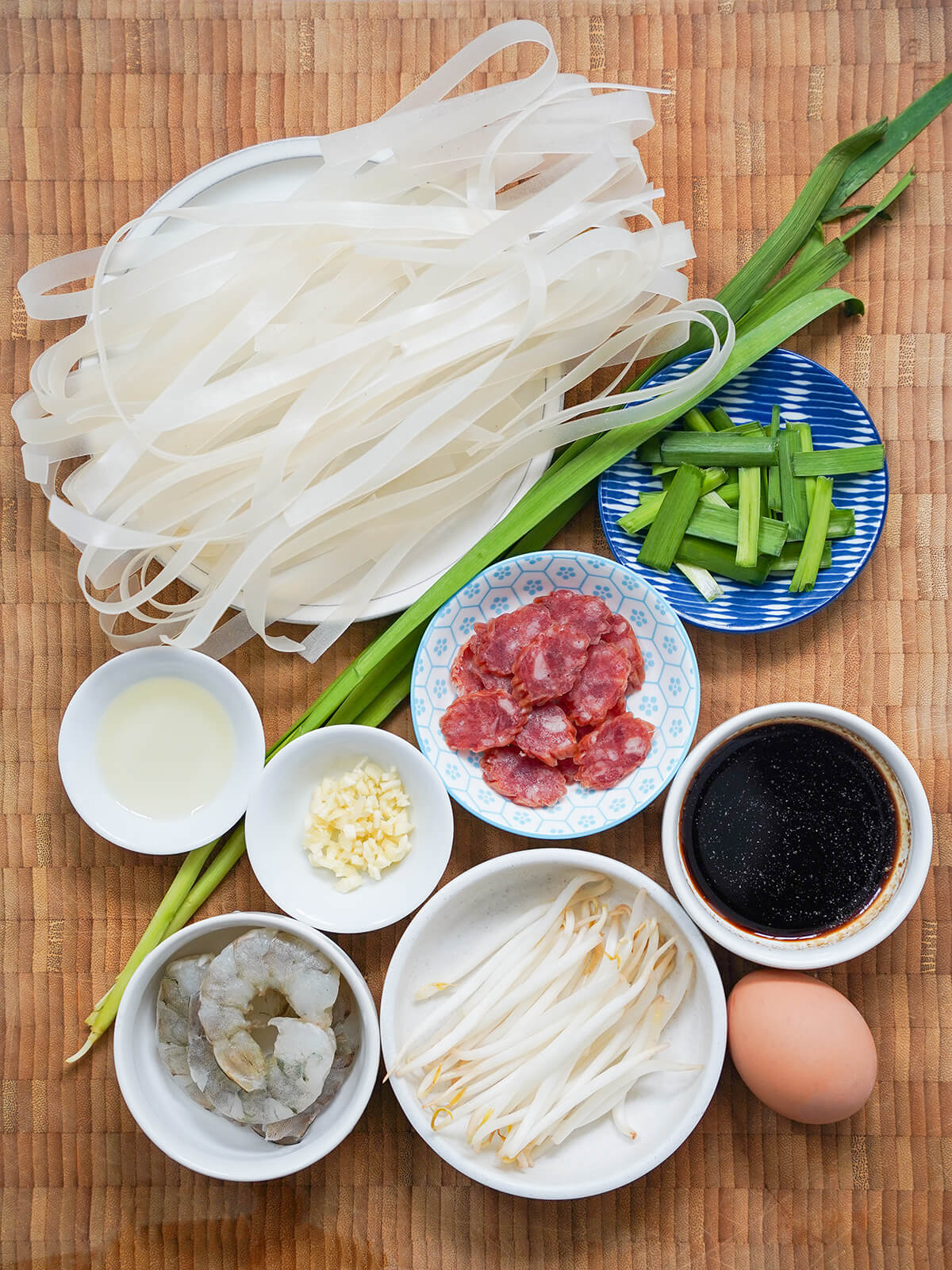 plate of rice noodles, small dishes with sliced sausage, garlic, chives, beansprouts, shrimp, sauce mixture and egg to side.