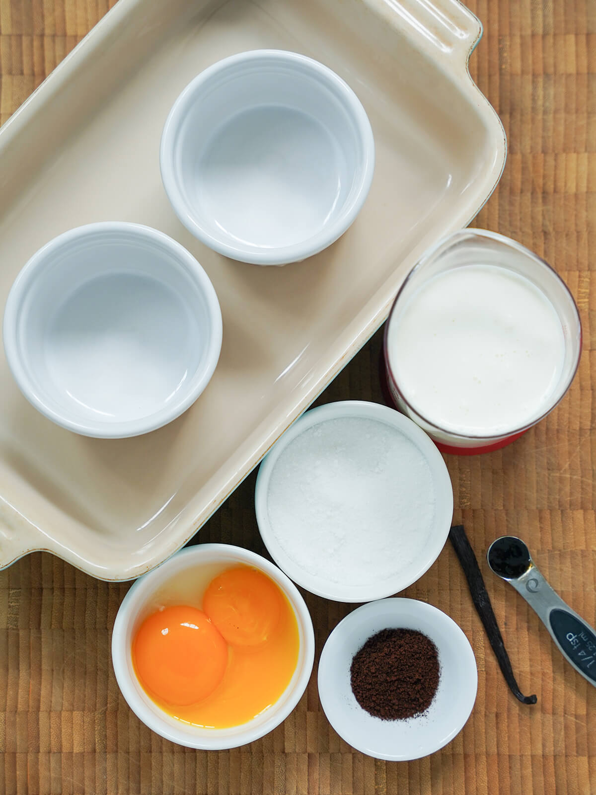 coffee creme brulee ingredients ramekins sitting in a baking dish with a measure of cream, bowl of sugar and bowl of egg yolks next to it with small dish with espresso powder and vanilla pod.