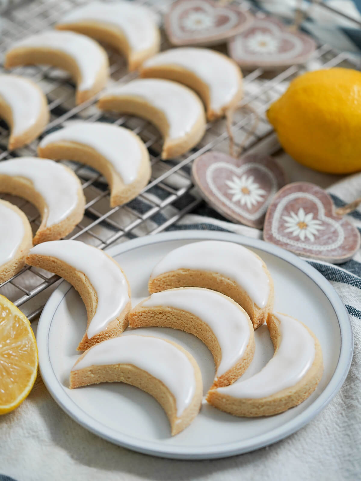 These lemon almond cookies (Zedernbrot) are so easy to make, with delicious light flavors and a wonderful chewy texture. Naturally gluten free, too, and with a bright lemon glaze, they're tasty little bites. plate of lemon almond cookies zedernbrot with more on cooling rack behind and wood decorations and lemons around.