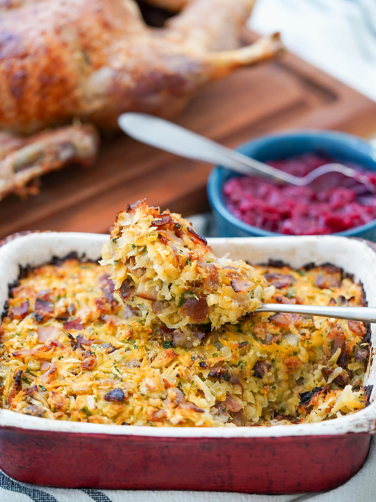 spoonful of rutabaga date and bacon stuffing on top of dish with rest, cranberry sauce and chopping board with duck blurred behind.
