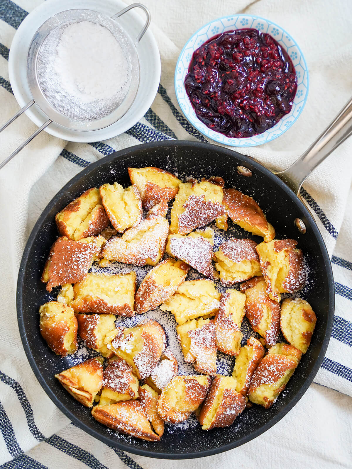 skillet of Kaiserschmarrn torn pancakes with dish of fruit compote and sift with powdered sugar above.