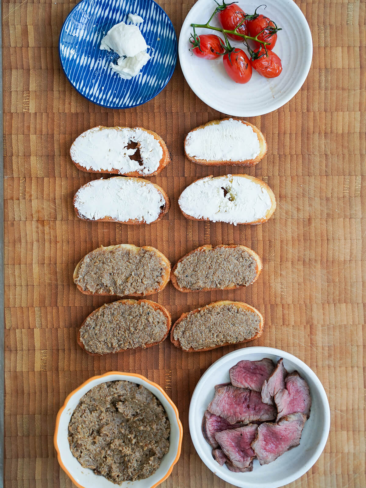 pieces of crostini, some topped with pate and others with goat cheese, with dishes above and below with pate, cheese, tomatoes and slices seared steak.