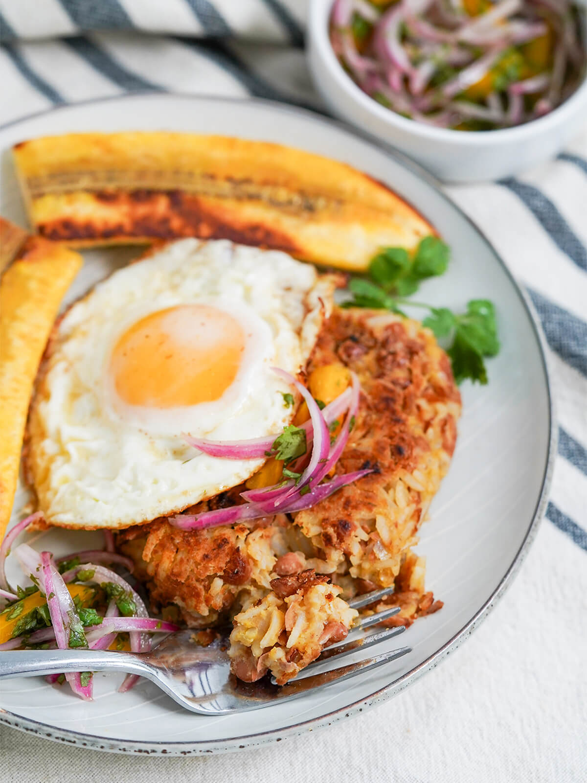 forkful of tacu tacu in front of rest of rice and bean cake topped with an egg, plantain behind.