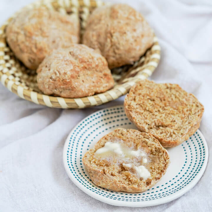 split open Irish soda bread roll with butter on it on plate in front of more rolls in basket behind.