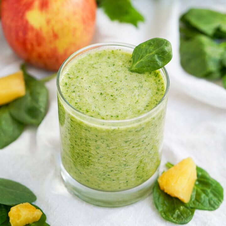 close view of glass of pineapple and spinach smoothie with pieces of pineapple and spinach leaves either side.
