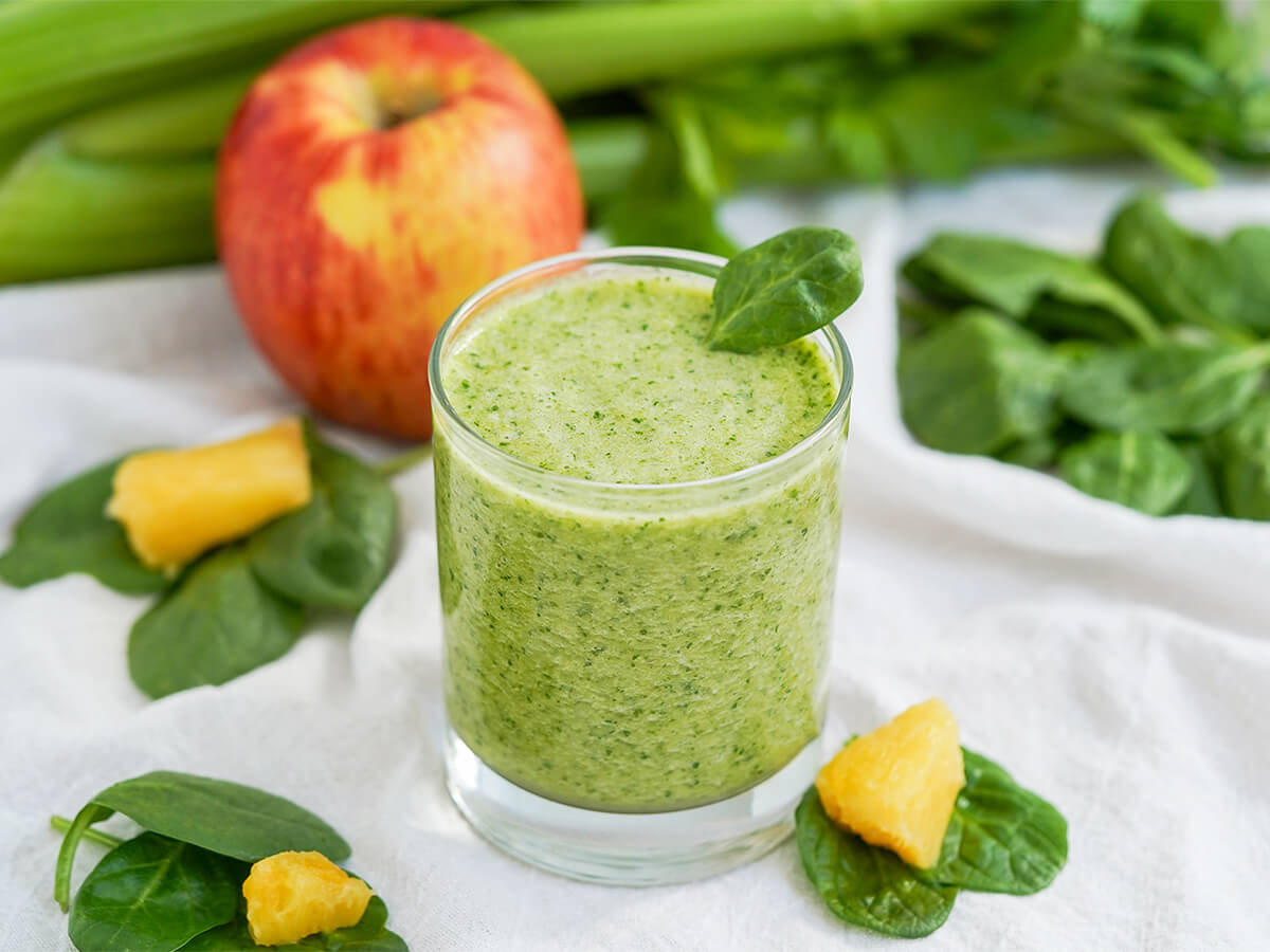 side view of glass of pineapple and spinach smoothie with spinach, pineapple chunks around glass and apple and celery behind.