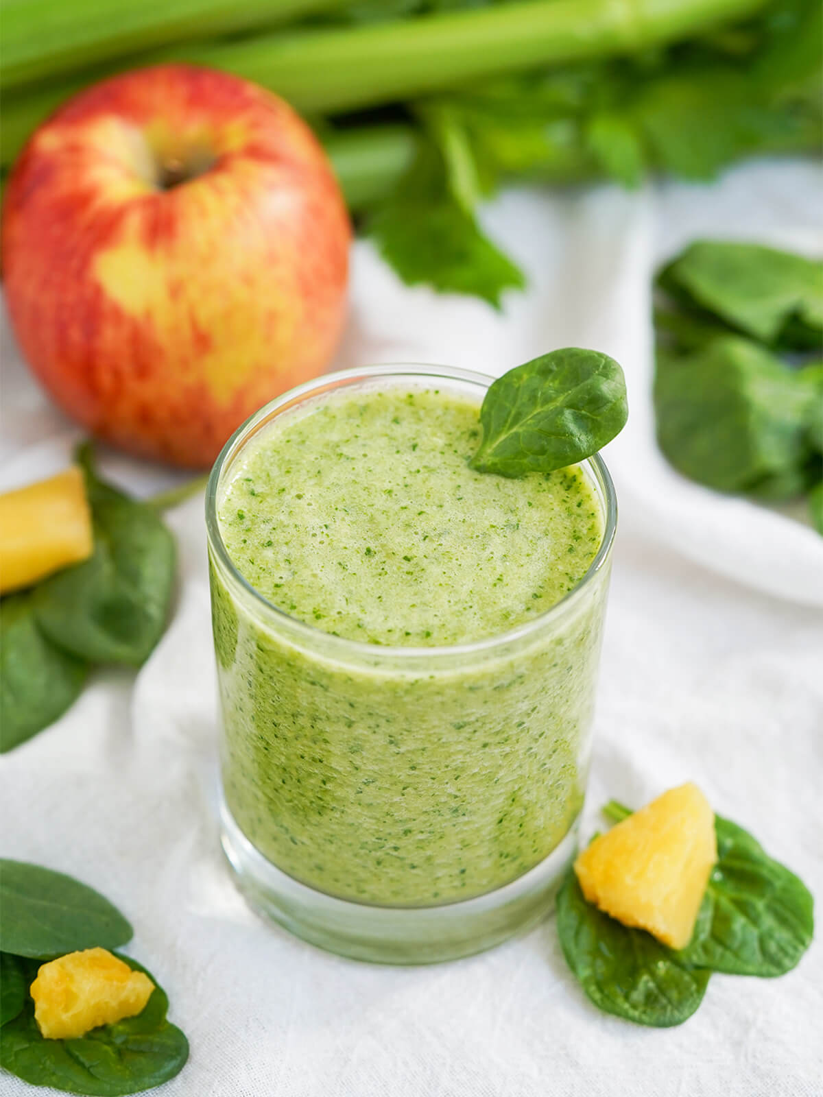 glass of pineapple and spinach smoothie with spinach leaves, pieces of pineapple around glass.