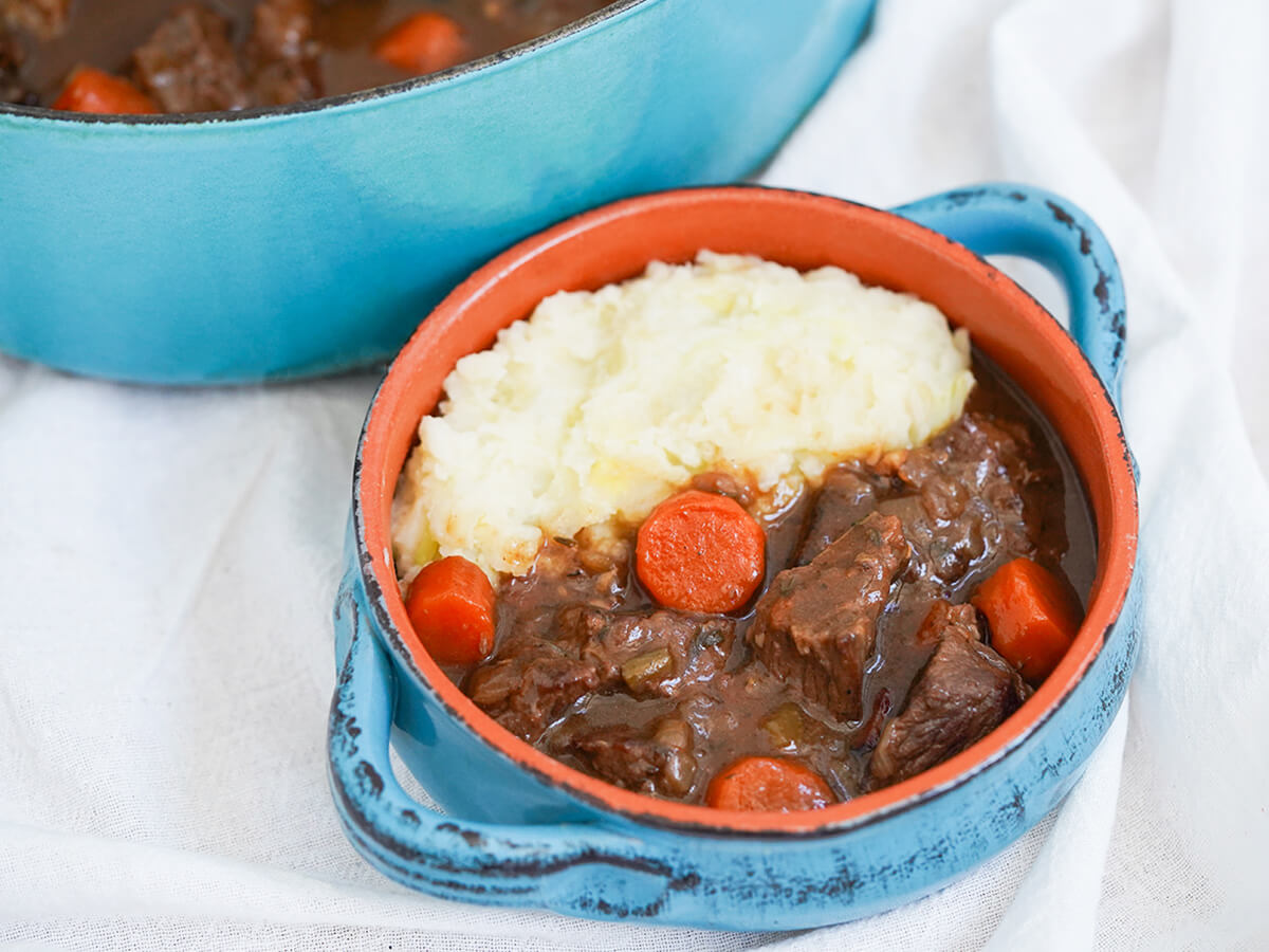 blue bowl of Guinness beef stew with mashed potato to side with pot in background.