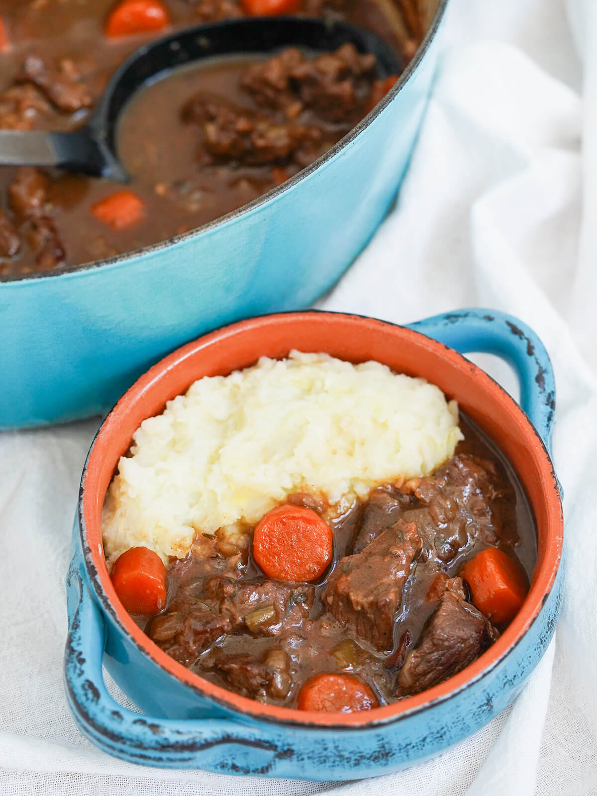 bowl of Guinness beef stew with mashed potato in back of bowl and pot with more stew and spoon in it in background.
