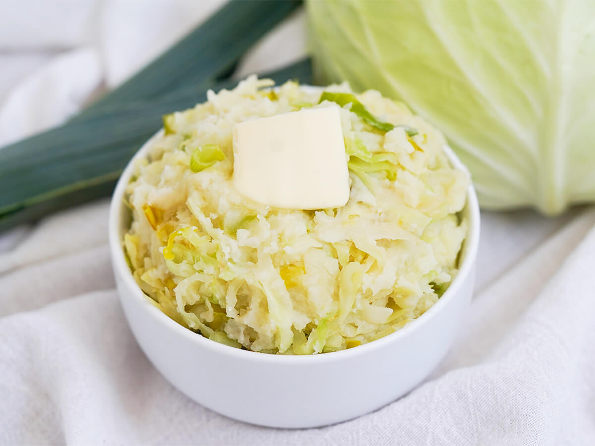 Colcannon is a simple side dish of mashed potatoes and cabbage that's just as good with leftovers as it is made fresh. Comforting, easy and great alongside so many dishes. bowl of colcannon, mashed potatoes with cabbage, with part cabbage and leek behind.