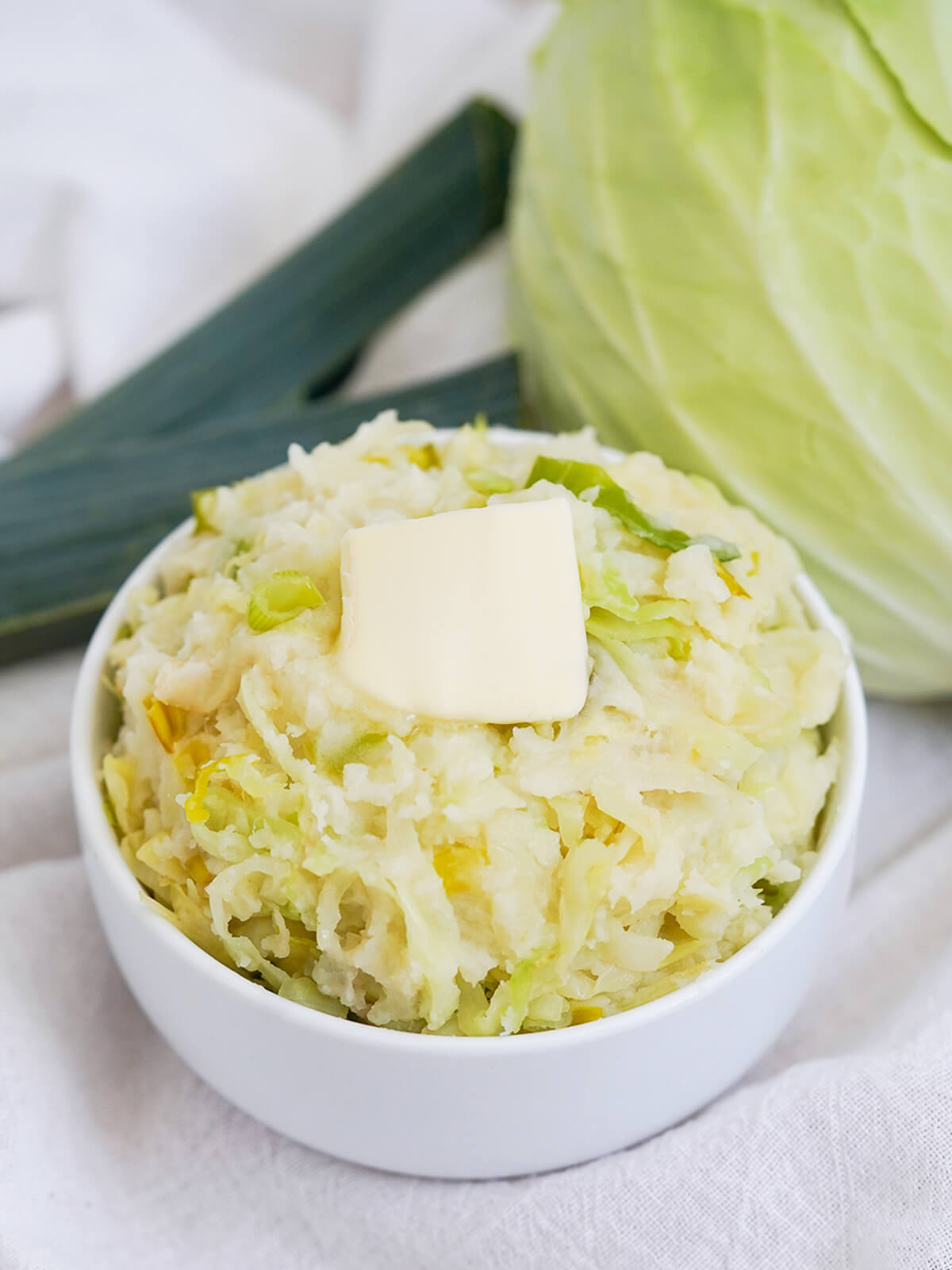 Colcannon is a simple side dish of mashed potatoes and cabbage that's just as good with leftovers as it is made fresh. Comforting, easy and great alongside so many dishes. bowl of colcannon, mashed potatoes with cabbage, topped with butter with cabbage and leek top in background behind.