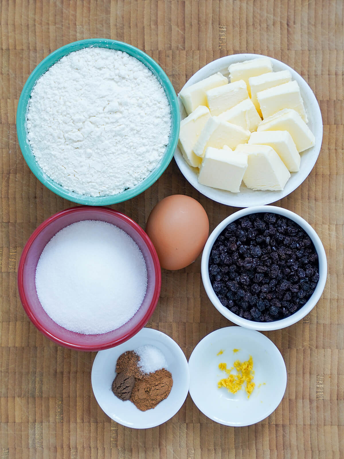 bowls of flour, sugar, currants, dish with chunks of butter and small dishes with spices, lemon zest and an egg in middle.